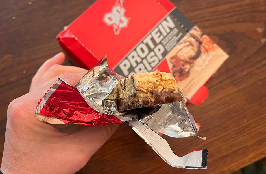 A hand holds a BSN Protein Crisp bar with a bite taken out. A box of bars can be seen in the background.