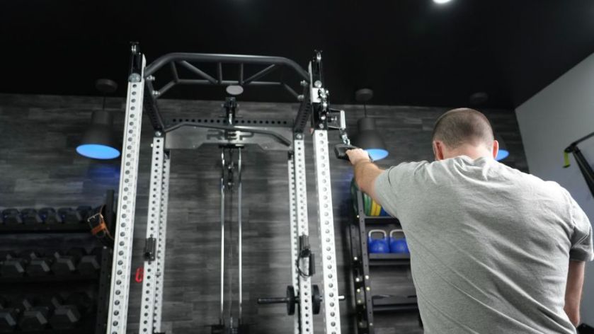 A person in a gray shirt using a cable machine in a gym. The gym has a dark ceiling with spotlights and shelves of dumbbells and blue kettlebells in the background.