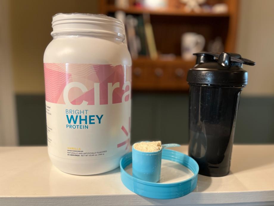 A scoop of Cira Bright Whey Protein powder is shown in front of the cntainer and a shake.