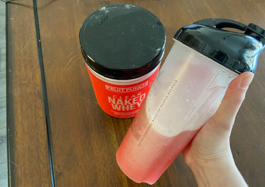 A hand holds up a shake of Clear Naked Whey Protein next to the container.