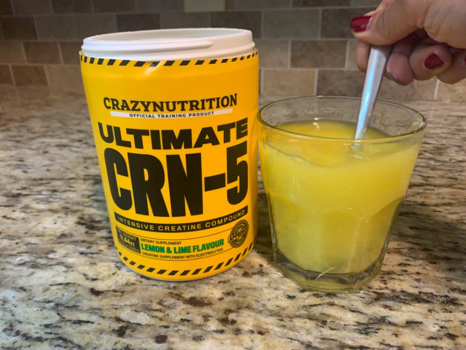 A hand is shown stirring Crazy Nutrition CRN-5 into a glass next to a yellow container.
