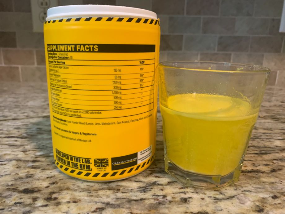 A very yellow drink in a glass rests next to a very yellow container of Crazy Nutrition Ultimate CRN-5 with the Supplement Facts label on display.