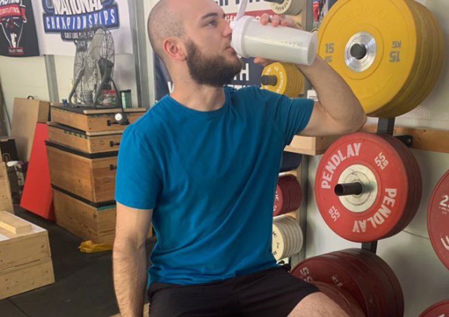 A man standing in a gym is drinking from a shaker bottle.