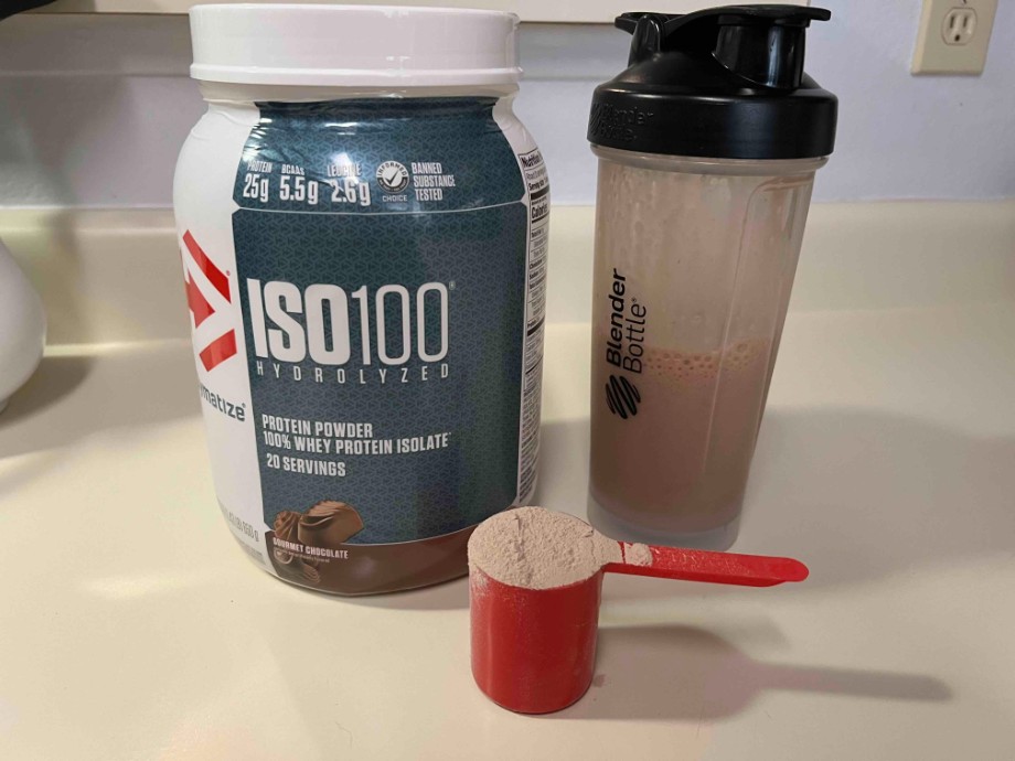 A container of A container and scoop of Dymatize ISO 100 next to a half-full shaker cup.