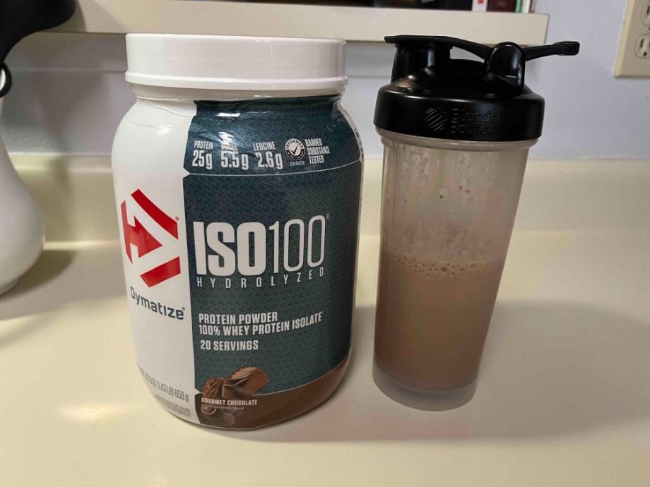 A container of Dymatize ISO 100 next to a fresh mixed shaker cup.