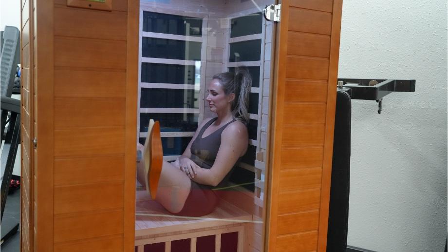 A woman sits inside a wooden infrared sauna, with her legs extended and arms resting on her knees. She appears relaxed and comfortable. The sauna is located in a modern room with a visible wall and some equipment in the background.
