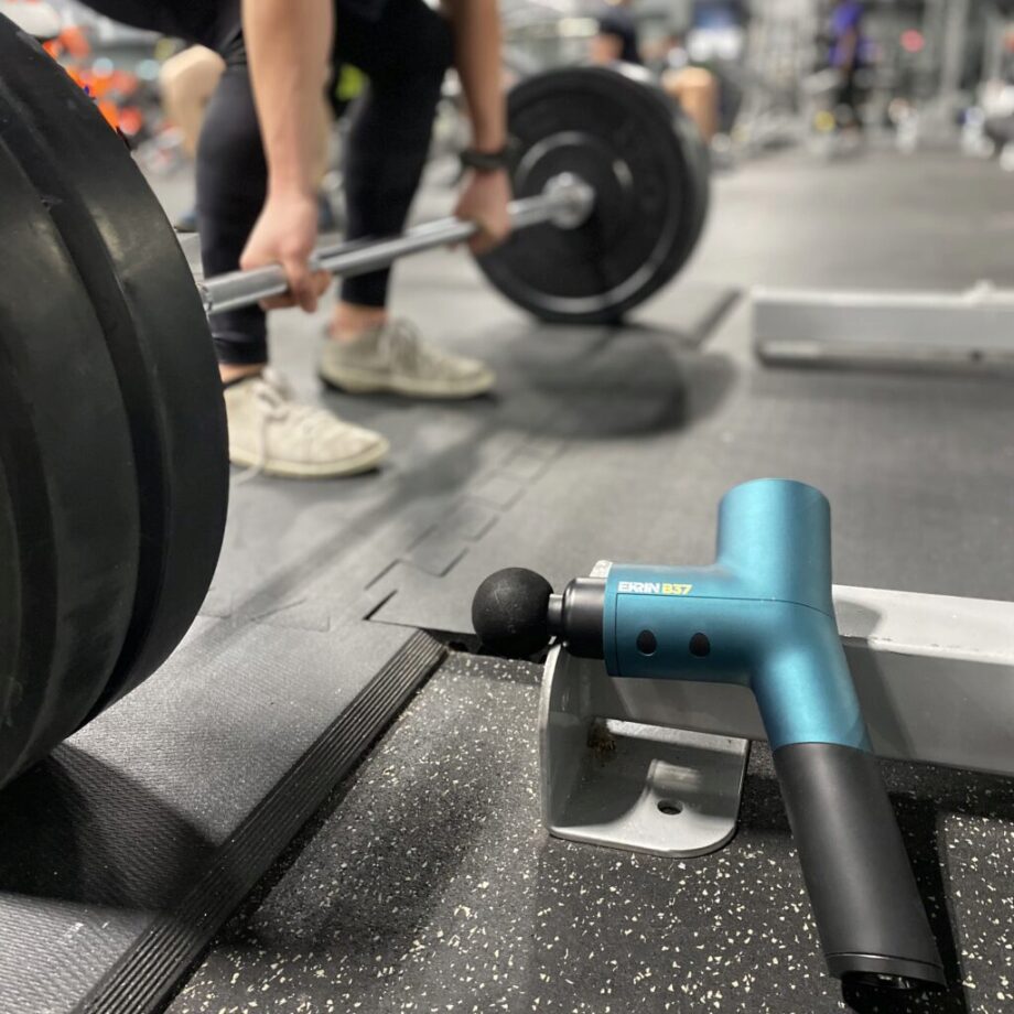 Ekrin Athletics b37 massage gun in focus while someone is doing deadlifts in the background