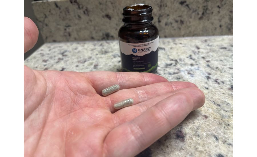 A hand holding two capsules filled with a greenish powder, in front of a brown bottle of Gnarly Nutrition Baseline Iron supplements.