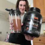 A smiling woman holds out a shake and a container of GNC AMP Wheybolic