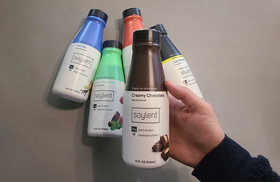 Someone holding a bottle of Soylent with bottles of different flavors in the background