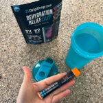 A hand holding a DripDrop ORS packet next to the package and a shaker bottle.
