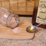 A person holds a spoon of honey near a jar of pink salt that's spilled on a wooden cutting board,