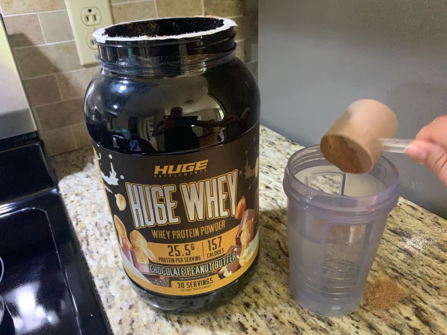 A scoop of Huge Whey Protein is being poured into a cup next to the prominently placed container