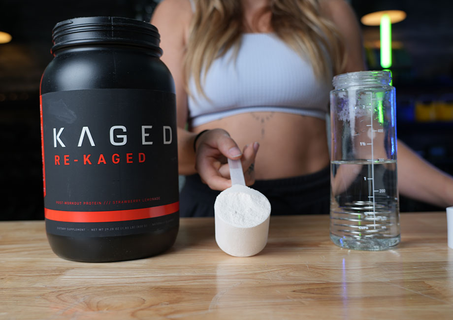 A person holds up a full scoop of Kaged re-Kaged between the container and a shaker glass of water.