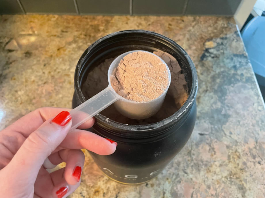 Looking down on a hand that's holding a scoop of Kaged Whey Protein Isolate above the container.