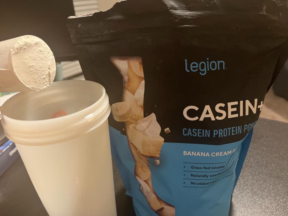 A bag of Legion Casein+ Protein Powder is next to a scoop being poured into a shaker cup.