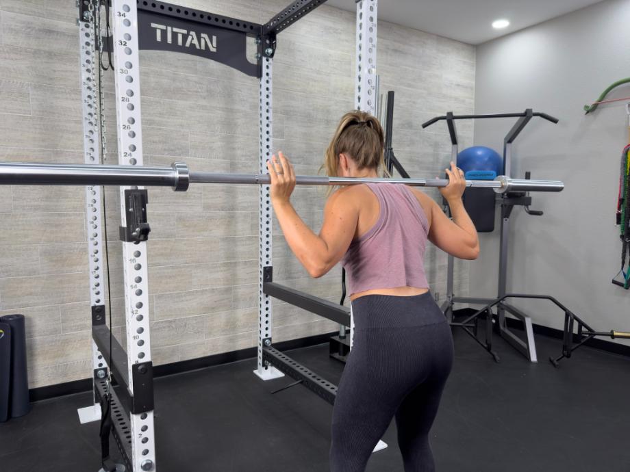 A woman stands holding a Living.Fit Barbell that is resting on her back