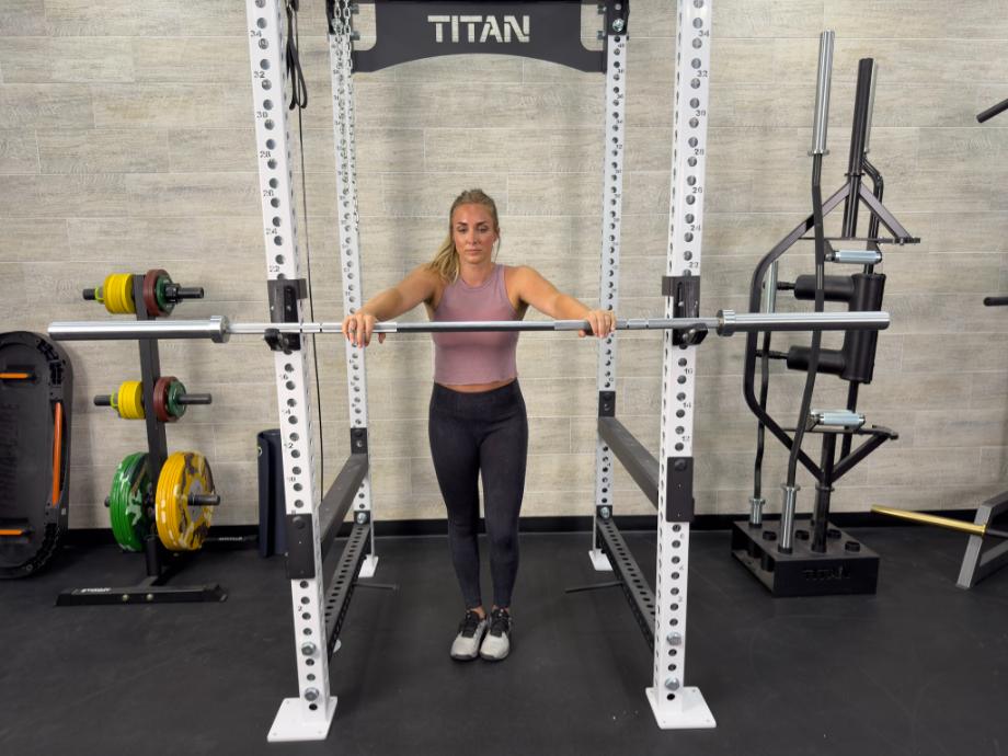 A woman is shown working out with the Living.Fit Barbell resting on a Power Rack