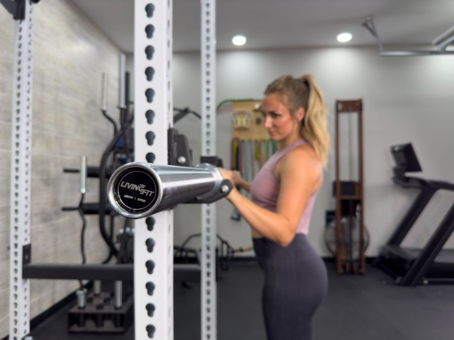 Woman standing with the Living.Fit Chrome Barbell placed in a squat rack