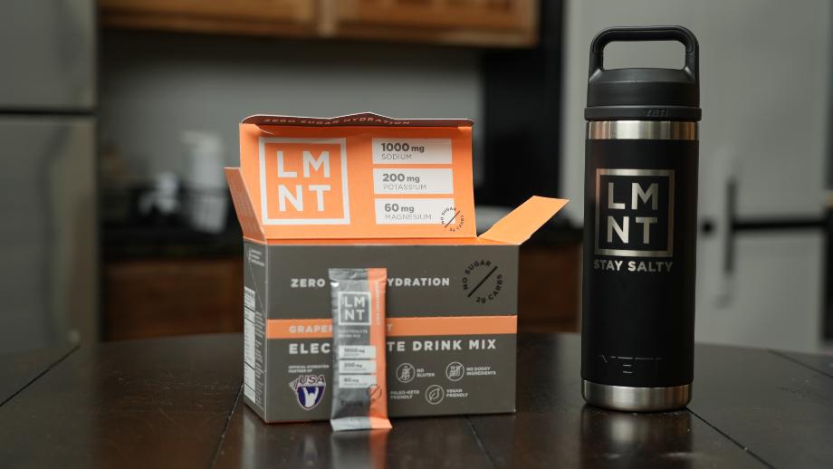 A packet of LMNT Electrolyte is leaning against the box, staged next to an LMNT shaker.