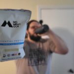 A man holds up a bag of Momentous Whey Protein while drinking a shake in the background.