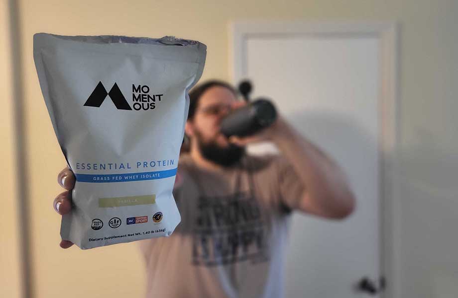 A man holds up a bag of Momentous Whey Protein while drinking a shake in the background.