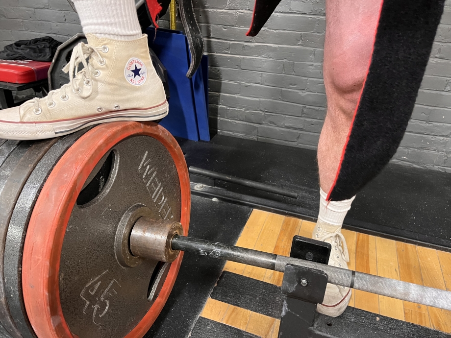 A person wearing Converse Chuck Taylor shoes steps on a barbell. 