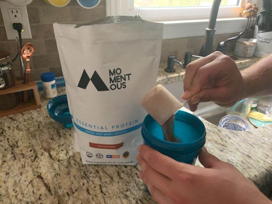 An open bag of Momentous Gras-Fed Whey Protein sitting on the counter while a hand pours a scoop of the product's fine brown chocolate powder into a shaker bottle.