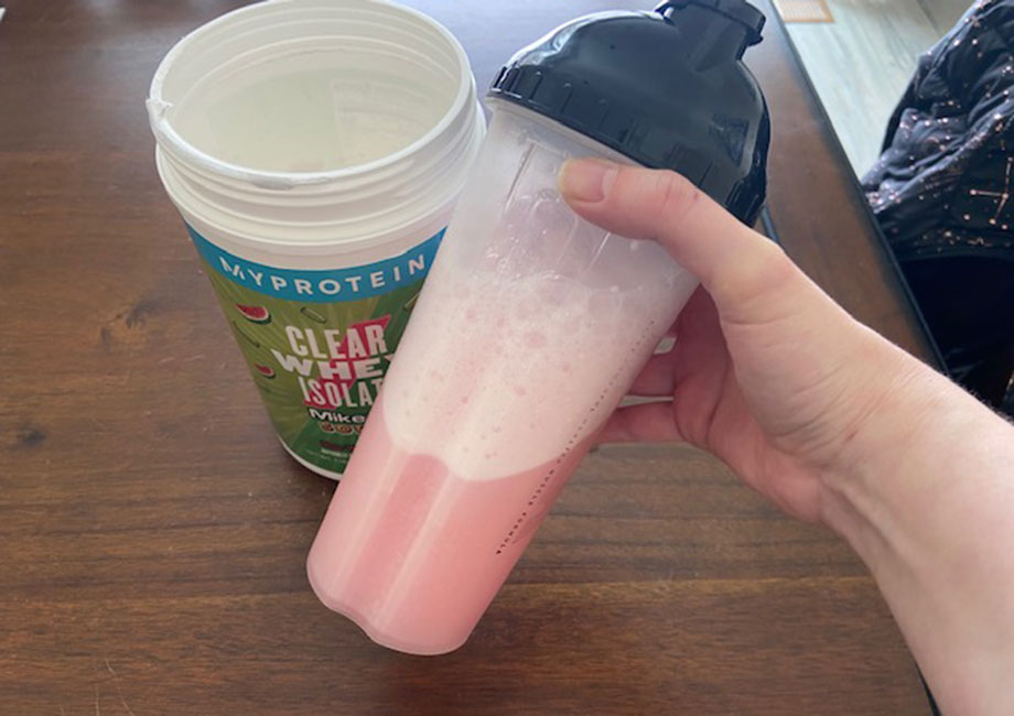 A hand holds out a shaker cup full of pink Myprotein Clear Whey Isolate.