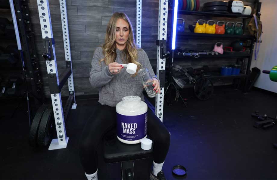 A woman is shown sitting on a weight bench, about to pour a comically large scoop of Naked Nutrition Naked Mass gainer into a reasonably sized shaker glass.