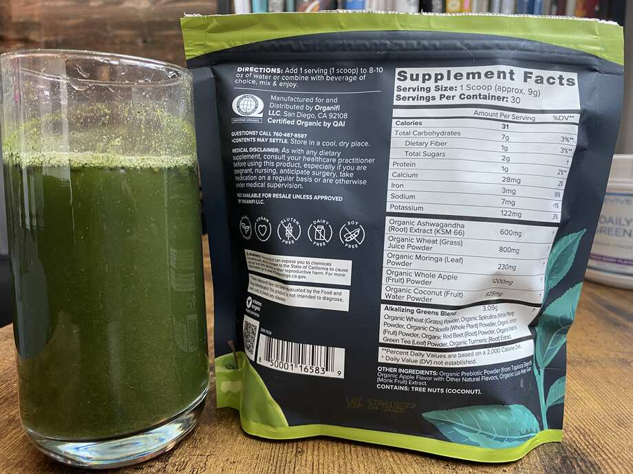 Close up of a tall glass of Organifi Greens Juice next to the bag with the Supplement Facts label showing.