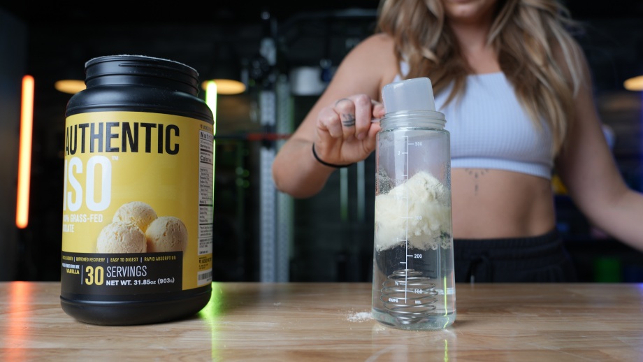 A person is shown dumping a scoop of Jacked Factory Authentic Iso Whey Protein powder into a shaker glass of water.