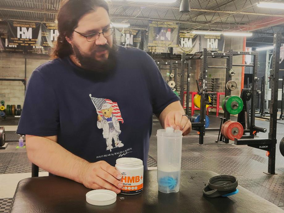 A man pours a scoop of Blonyx HMB+ Creatine into a shaker cup while inside of a gym