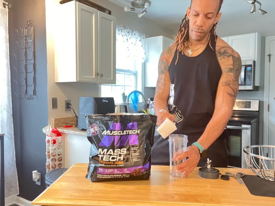 A person stands in a kitchen pouring a scoop of MuscleTech Mass Gainer into a shaker cup.