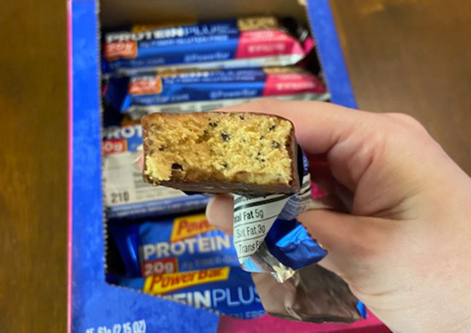 A hand holds a PowerBar Protein Plus bar with a bite taken out of it.