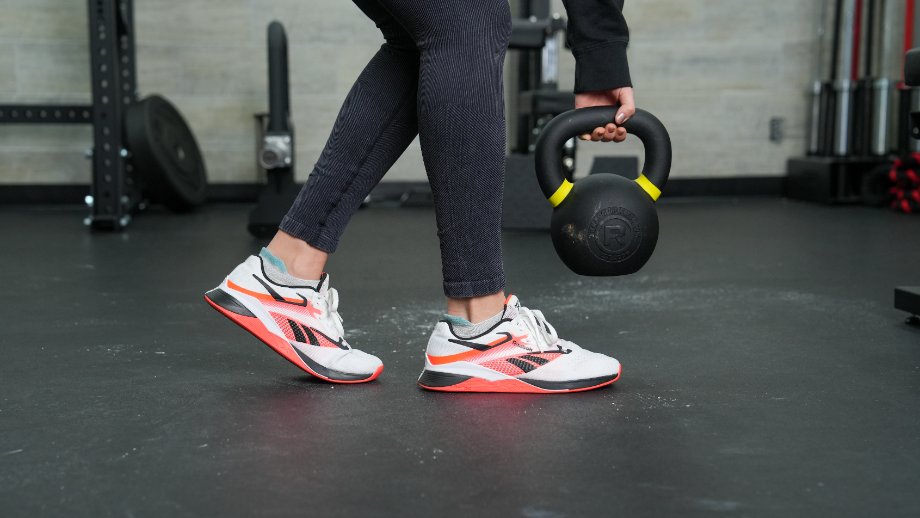 A person is shown walking in Reebok Nano X4 shoes while carrying a kettlebell.