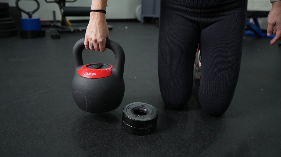 A REP Fitness Adjustable Kettlebell is held next to a weight plate.