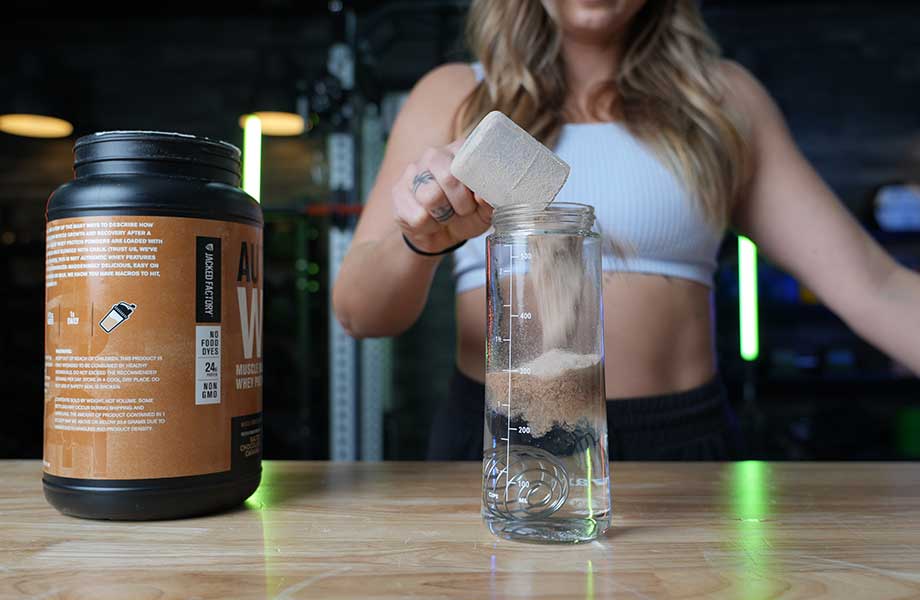 A woman is shown pouring a scoop of Jacked Factory Authentic Whey into a shaker glass.