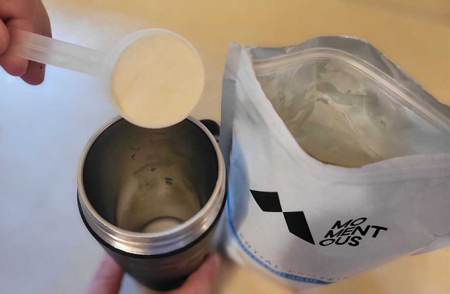 Close up view looking down on a hand scooping Momentous Grass-Fed Whey Protein into a stainless steel mug.