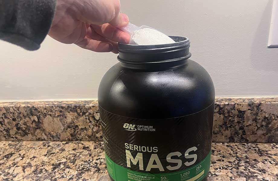A hand carefully lifts a scoop of powder from a container of Optimum Nutrition Serious Mass