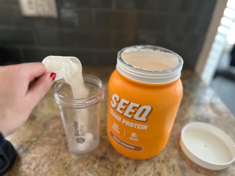 A scoop of SEEQ Clear Whey Protein Isolate is being poured into a shaker cup.