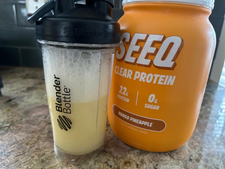 A freshly mixed shaker bottle of SEEQ Clear Whey Protein Isolate is creamy and foamy looking next to the SEEQ canister