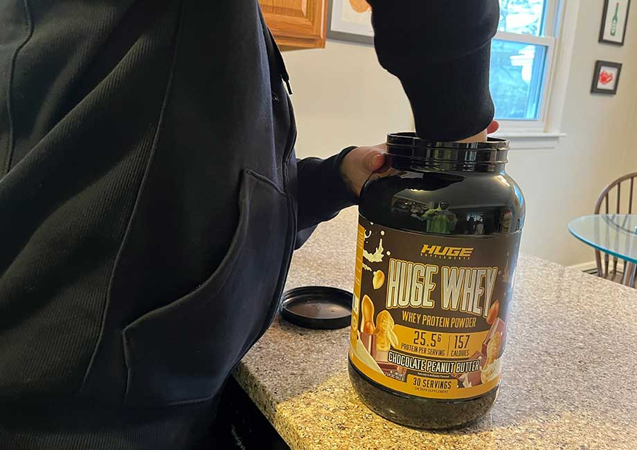 A person reaching down into a container of Huge Whey Protein to scoop some out