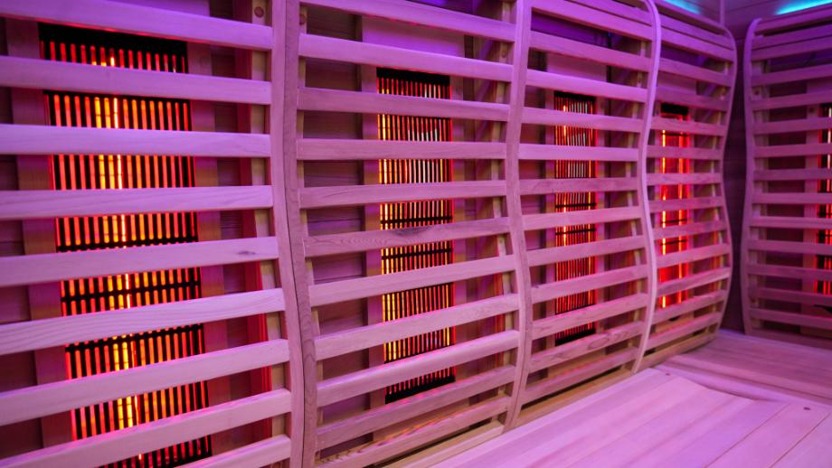 Inside view of an infrared sauna featuring wooden slatted walls with glowing red heating elements. The ambient lighting is purple, creating a warm and relaxing atmosphere.