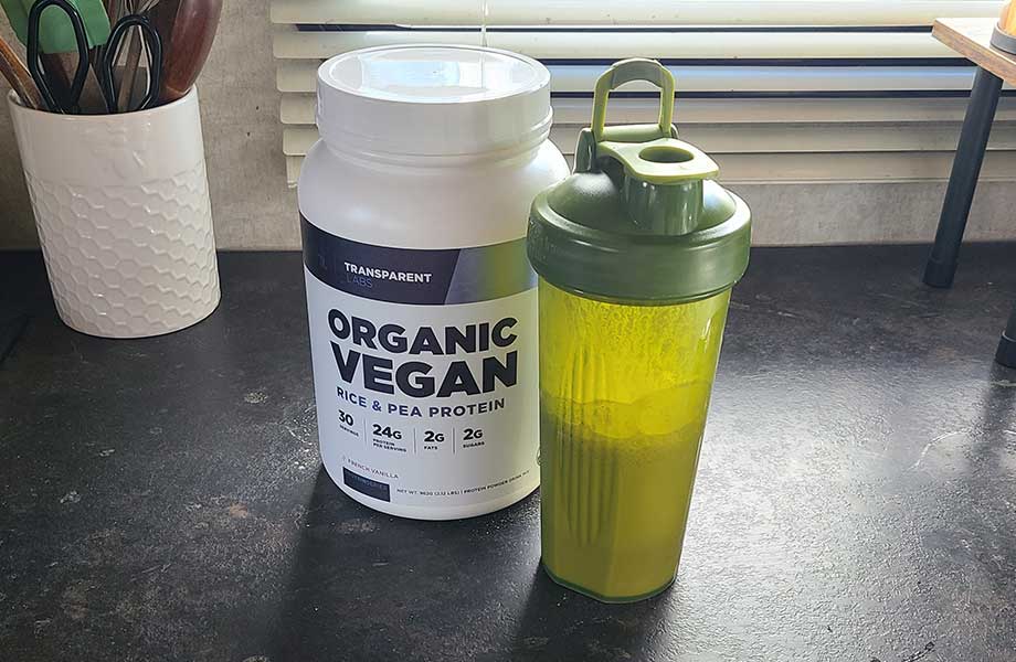 A half-full green shaker bottle rests next to a container of Transparent Labs Vegan Protein powder