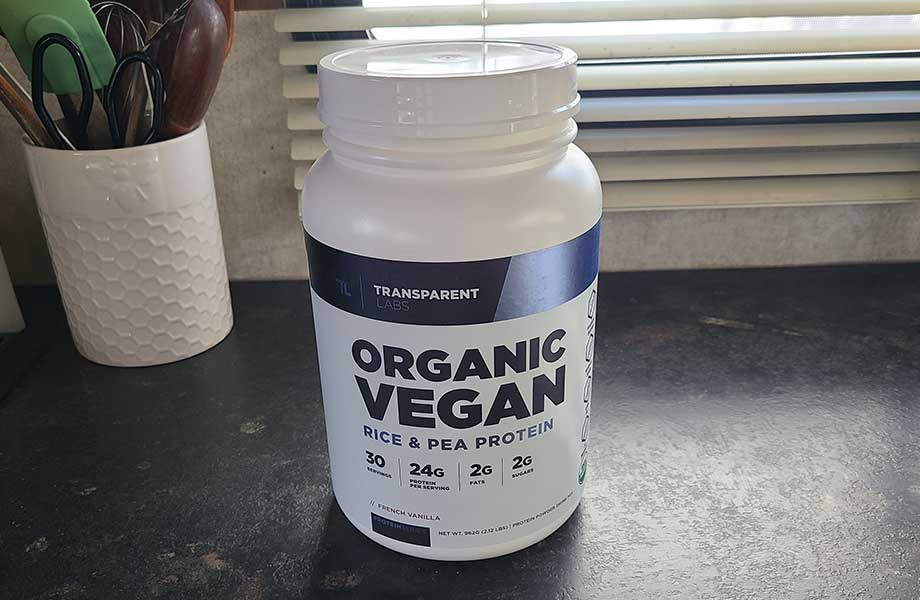 A container of Transparent Labs Vegan Protein powder sits on a counter, and a cup of random kitchen stuff can be seen in the background.