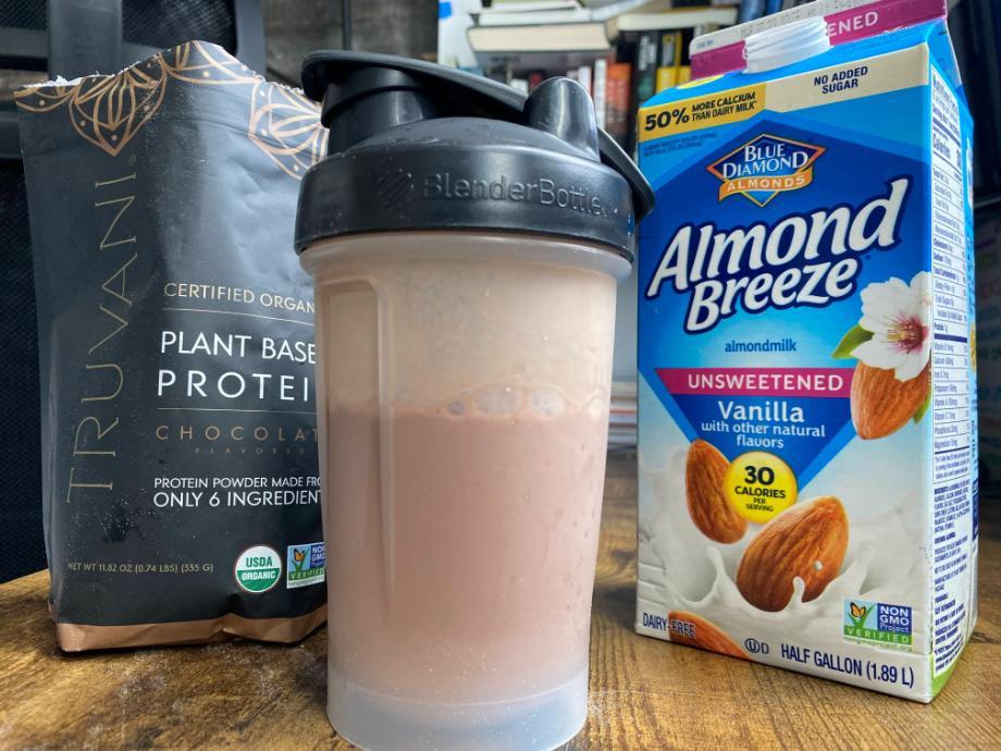 A Truvani Chocolate Protein shake stands in front of a bag of Truvani and a carton of almond milk