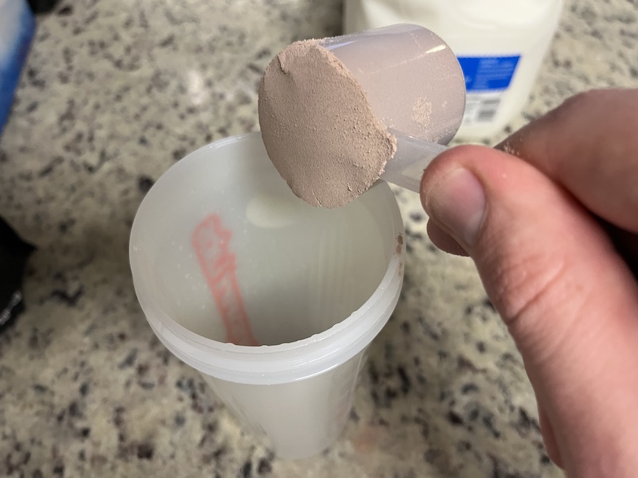 Close-up of a hand pouring a scoop of XWERKS Grow's fine-grained chocolate protein powder into a shaker bottle with an unfocused carton of milk sitting in the background.